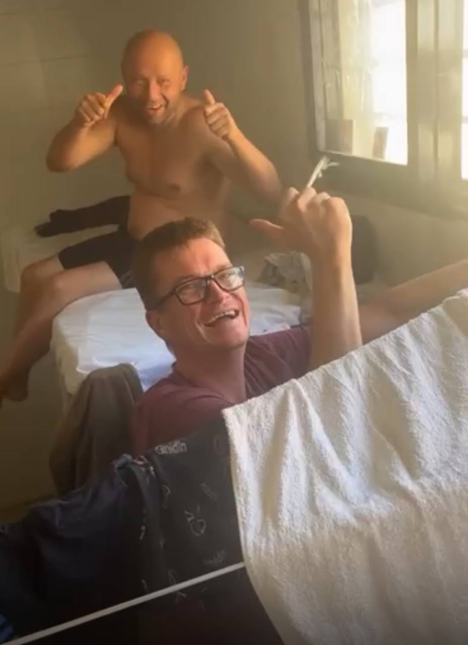 Two grinning inmates relaxing on a bunk in their prison cell, one giving double thumbs up