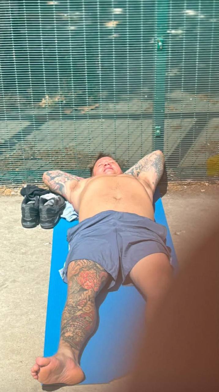 A tattooed inmate sunbathing on a blue yoga mat beside the prison perimeter fence